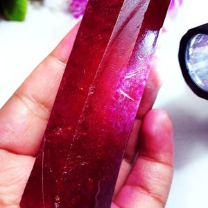 May include: A large, elongated, raw ruby crystal with a deep, saturated red colour and a translucent, faceted-like texture. The stone displays natural surface striations and internal inclusions, catching light to reveal varying shades of crimson.