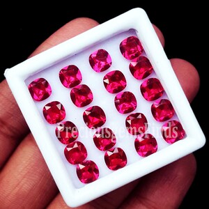 May include: A collection of eighteen vibrant, cushion-cut red gemstones arranged in a grid within a white display box. Each stone features a faceted surface that reflects light, creating a brilliant appearance. The text Preciousgemsfinds is visible.
