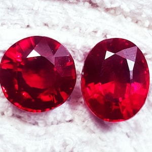 May include: Two faceted gemstones with a deep red colour. One stone is circular in shape, while the other is oval. Both gems feature intricate geometric cuts that reflect light, creating a brilliant appearance across their polished surfaces.