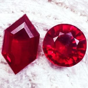 May include: Two faceted, deep red gemstones. One is a hexagonal shape, the other is round. Both have multiple facets that reflect light, creating a brilliant sparkle. The gems are set against a white, textured background.