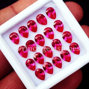 Peut inclure: Une boîte de présentation rectangulaire blanche contenant vingt pierres précieuses rouges à facettes en forme de poire, disposées en grille. Les pierres présentent une couleur rouge vif et profond avec des surfaces réfléchissantes. Le texte "prince gemstones" est imprimé en blanc.