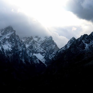 May include: A dramatic view of snow-capped mountain peaks with a bright, white sky and dark, stormy clouds.
