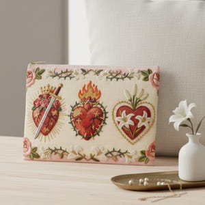 May include: Cream-colored embroidered pouch featuring three sacred heart designs with floral and crown details. The pouch has a pink border with floral and thorn accents, and a gold zipper.