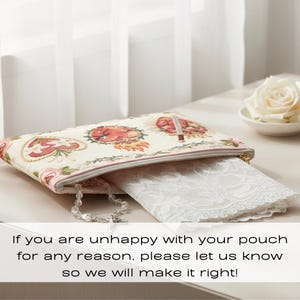 May include: A cream-colored pouch with a floral and heart design, a pink zipper, and a gold zipper pull. A white lace item is partially visible inside. A rosary and a white rose are also present.