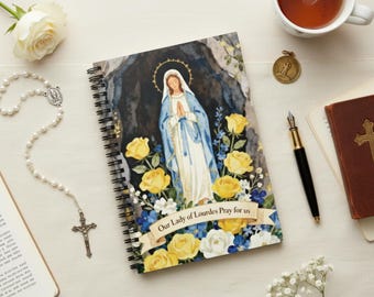 May include: A spiral-bound notebook featuring an illustration of Our Lady of Lourdes, surrounded by yellow roses and blue flowers. The text "Our Lady of Lourdes Pray for us" is on a banner. A rosary, a cup of tea, and a pen are also visible.