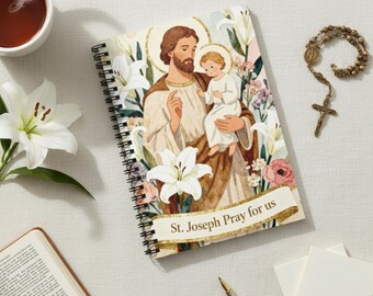 May include: A spiral-bound notebook featuring an illustration of St. Joseph holding a child, surrounded by lilies and flowers. The text "St. Joseph Pray for us" is on a banner. A rosary, open book, pen, and cup of tea are also visible.