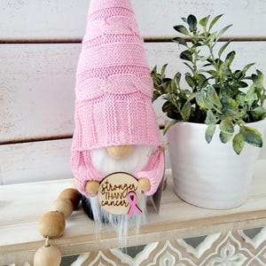 Gnome with a white beard and a pink knitted hat holding a wooden sign that says "stronger than cancer" with a pink ribbon shown displayed on a shelf next to a plant.