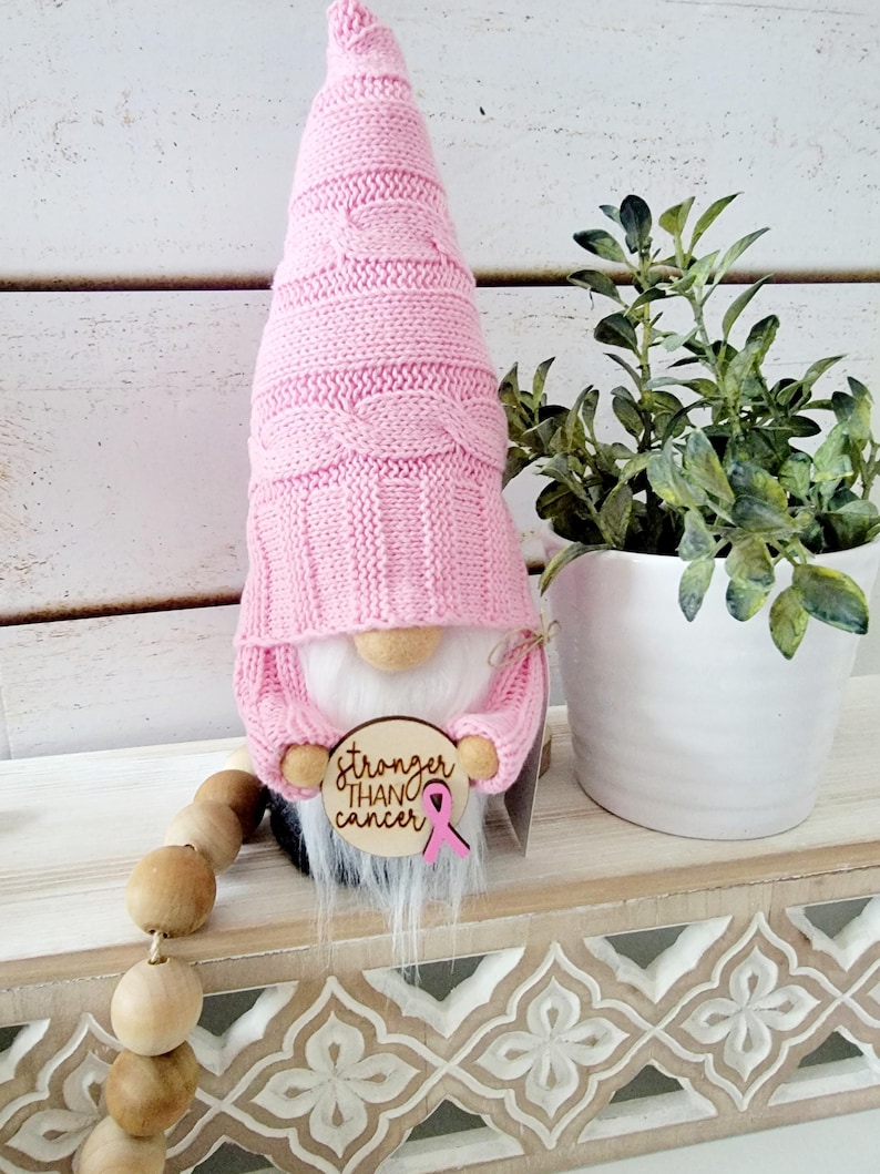 Gnome with a white beard and a pink knitted hat holding a wooden sign that says "stronger than cancer" with a pink ribbon shown displayed on a shelf next to a plant.