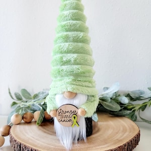 May include: A green plush gnome holding a wooden sign with the text "Stronger Than Cancer" and a yellow ribbon.