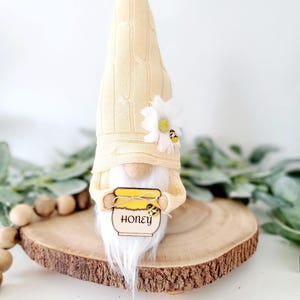 May include: A whimsical gnome figurine with a cream-colored knit hat and a long white beard. The gnome holds a wooden sign that reads "HONEY" and features a bee illustration. A white flower with a yellow center and a bee accent the hat. The figurine rests on a wooden slice.