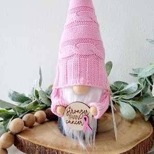 A pink gnome holding a wooden circle with the text "stronger than cancer" engravrd and a pink ribbon embellishment attached to the sign.