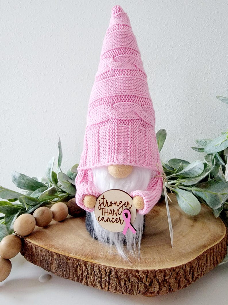A pink gnome holding a wooden circle with the text "stronger than cancer" engravrd and a pink ribbon embellishment attached to the sign.