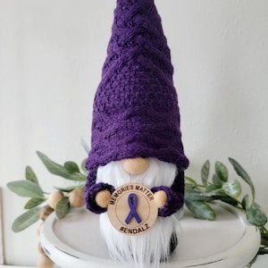 May include: A handmade gnome with a purple knit hat and beard. The gnome holds a wooden sign with a purple ribbon and the words "Memories Matter" and "#ENDALZ". The gnome is on a white decorative stand.