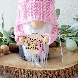 A pink gnome holding a wooden circle with the text "stronger than cancer" engravrd and a pink ribbon embellishment attached to the sign.