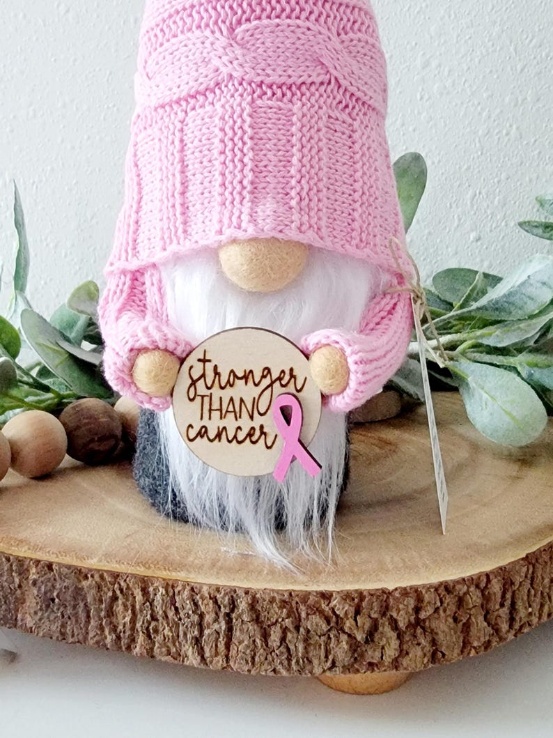 A pink gnome holding a wooden circle with the text "stronger than cancer" engravrd and a pink ribbon embellishment attached to the sign.