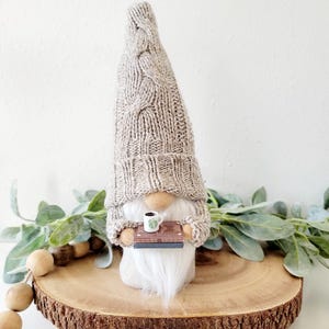 May include: A handcrafted gnome figurine with a long, gray knit cap and matching sweater. The gnome has a white beard and is holding a small wooden tray with a miniature coffee cup. The figurine is displayed on a wooden slice.