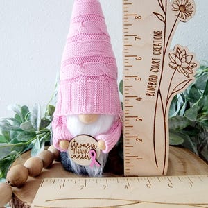 A pink gnome holding a wooden sign that says "Stronger Than Cancer" with a pink ribbon. The gnome is standing next to a wooden ruler showing 4" wide by 10" tall