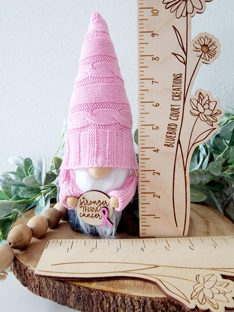 A pink gnome holding a wooden sign that says "Stronger Than Cancer" with a pink ribbon. The gnome is standing next to a wooden ruler showing 4" wide by 10" tall