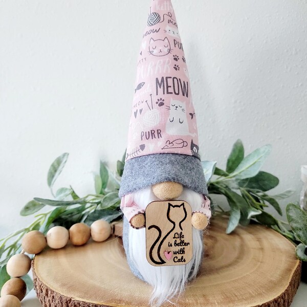 May include: A cat-themed gnome figurine with a pink and gray pointed hat. The hat is patterned with cat faces, yarn, and the words "MEOW" and "PURR". The gnome holds a wooden sign that reads "Life is better with Cats".