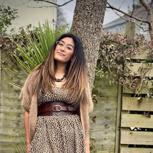 May include: A woman wearing a brown and black leopard print dress, a brown belt, and a tan sweater. The dress has a pleated skirt. The belt is wide with a large silver buckle. She is wearing glasses and a black necklace.