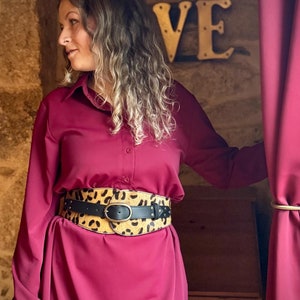 May include: A burgundy dress with a wide belt featuring a leopard print design and a black leather strap. The belt has a large oval buckle and is cinched at the waist. The dress has long sleeves and a button-down front.