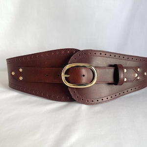 May include: A wide, dark brown leather belt with a gold-colored oval buckle. The belt has decorative stitching and small gold-colored studs. The design is a wide corset style.