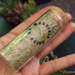 May include: A glass jar with a cork lid filled with dried herbs. The jar has a black label that reads "Mountain Spice Co."