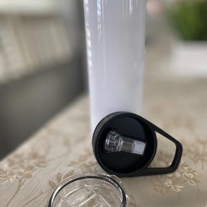 May include: A white, cylindrical tumbler with a black lid and a clear, open lid. The black lid has a handle and a clear plastic spout. The tumbler is designed to keep beverages cold or hot.
