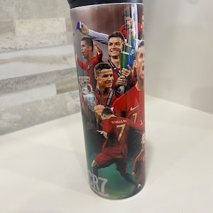 May include: A stainless steel tumbler with a black lid and handle. The tumbler features a collage of images of Cristiano Ronaldo, including photos of him playing soccer, holding trophies, and celebrating. The text "CR7" and "RONALDO" are visible on the tumbler.