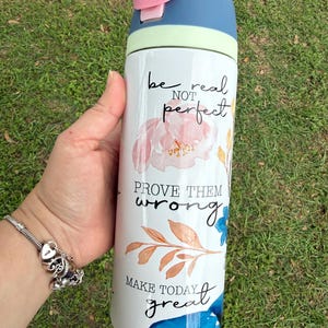 K&ouml;nnte beinhalten: Eine wei&szlig;e Wasserflasche mit blauem und rosa Deckel, verziert mit floralen Mustern und den S&auml;tzen &bdquo;be real not perfect&ldquo;, &bdquo;prove them wrong&ldquo; und &bdquo;make today great&ldquo;. Die Flasche wird vor einem Hintergrund aus gr&uuml;nem Gras gehalten.