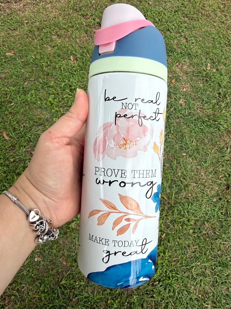 K&ouml;nnte beinhalten: Eine wei&szlig;e Wasserflasche mit blauem und rosa Deckel, verziert mit floralen Mustern und den S&auml;tzen &bdquo;be real not perfect&ldquo;, &bdquo;prove them wrong&ldquo; und &bdquo;make today great&ldquo;. Die Flasche wird vor einem Hintergrund aus gr&uuml;nem Gras gehalten.