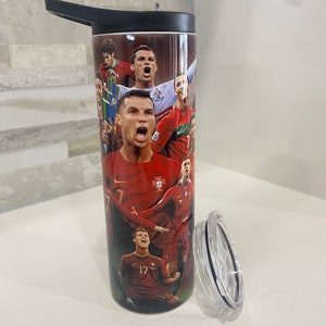 May include: A stainless steel tumbler with a black lid and a black handle. The tumbler features a collage of images of Portuguese soccer players, including Cristiano Ronaldo, wearing the red and green Portuguese national team jersey. The tumbler has a clear plastic lid with a black ring around the edge.