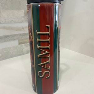 May include: A red, green, and gold striped stainless steel tumbler with a black lid and a black straw. The tumbler has the word "SAMII" printed on it.