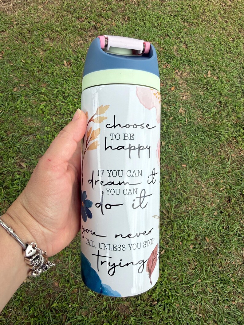 K&ouml;nnte beinhalten: Eine wei&szlig;e Wasserflasche mit blauem, gr&uuml;nem und rosa Deckel. Die Flasche ist mit floralen Designs und den Worten "choose to be happy", "if you can dream it, you can do it" und "you never fail, unless you stop trying" verziert.
