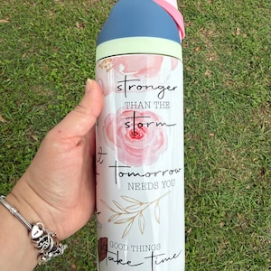 K&ouml;nnte beinhalten: Eine wei&szlig;e Wasserflasche mit blauem und rosa Deckel, mit dem Text "stronger than the storm" und anderen inspirierenden Phrasen. Die Flasche hat ein Blumendesign mit rosa Rosen und goldenen Akzenten.