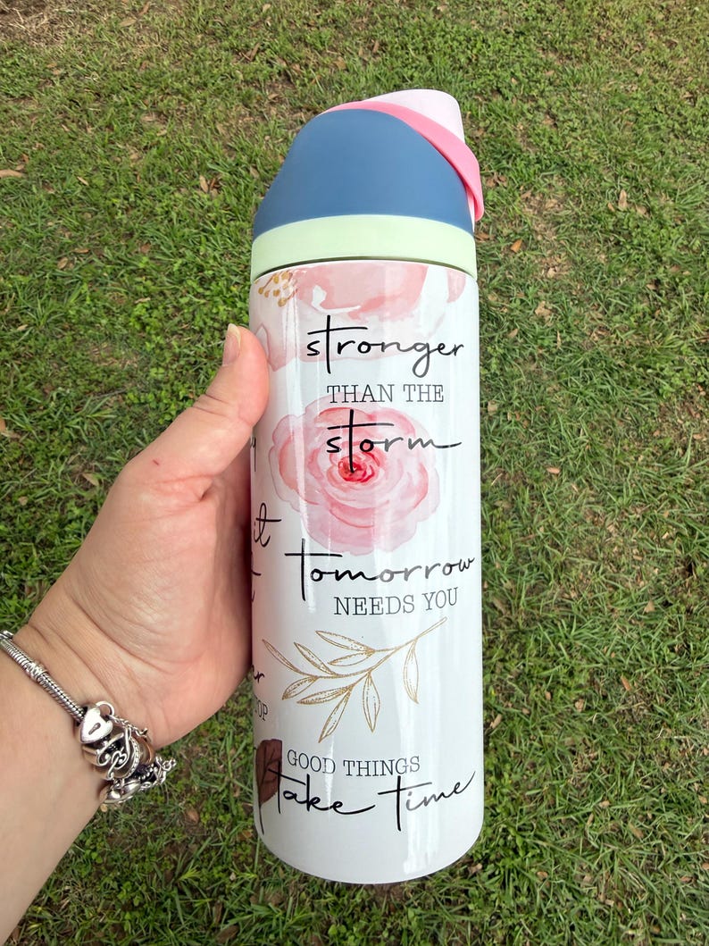 K&ouml;nnte beinhalten: Eine wei&szlig;e Wasserflasche mit blauem und rosa Deckel, mit dem Text "stronger than the storm" und anderen inspirierenden Phrasen. Die Flasche hat ein Blumendesign mit rosa Rosen und goldenen Akzenten.