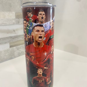 May include: A stainless steel tumbler with a red, white, and green design featuring multiple images of soccer players in Portugal's national team uniform. The tumbler has a black lid and a white base.