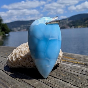Puede incluir: Un colgante azul claro en forma de lágrima con una fianza plateada. La piedra tiene un aspecto veteado con patrones blancos y azules más oscuros. El colgante descansa sobre un trozo de coral contra un fondo de lago y montañas.