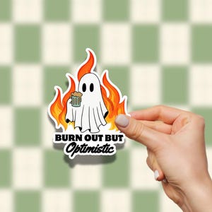 May include: A white ghost sticker with a cartoon design, holding a beer mug while surrounded by flames. The sticker has the text "BURN OUT BUT Optimistic" in black and white.