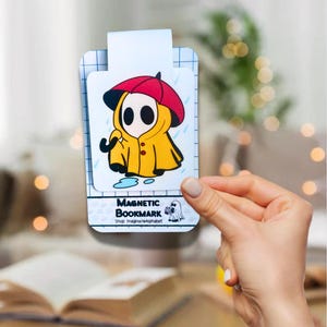 Peut inclure: Un marque-page magnétique avec un personnage de dessin animé portant un imperméable jaune et un parapluie rouge. Le personnage se tient dans une flaque d'eau. Le marque-page est blanc avec des accents bleus et le texte "Magnetic Bookmark" et "Shop: ImagineAlphabet".