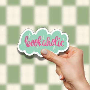 Peut inclure: Autocollant en forme de nuage vert clair avec le mot "bookaholic" en rose. Le texte est orné d'étoiles blanches. L'autocollant est tenu devant un fond à carreaux verts et blancs.