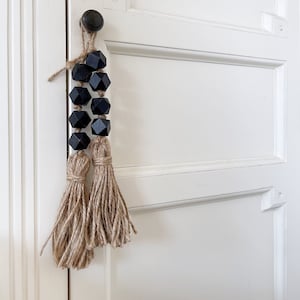 Peut inclure: Deux glands en bois noir avec une frange en jute suspendus à une porte blanche.