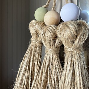 May include: Three macrame tassels with wooden beads in green, beige, and light blue colors. The tassels are made of natural jute fiber.