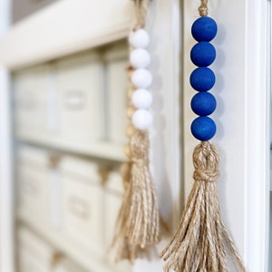 May include: A blue and white beaded garland with a jute tassel. The garland is made of wooden beads and has a rustic, farmhouse style.