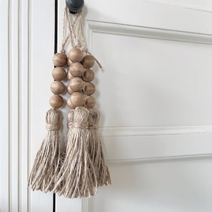 Peut inclure: Trois glands de perles en bois avec une frange en jute naturelle suspendus à un bouton de porte blanc.