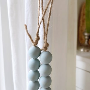 Puede incluir: Una cadena de cuentas de madera de color azul claro cuelga de un cord&oacute;n de cuerda natural. Las cuentas son redondas y lisas, y la cuerda est&aacute; anudada en la parte superior.