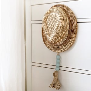 Op de afbeelding: Een strooien fedora hoed met een geweven patroon hangt aan een witte ladekast. Een decoratieve kwast met lichtblauwe kralen en een kwast van natuurlijke vezels is aan de hoed bevestigd.
