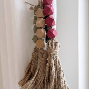 May include: A decorative beaded tassel with a mix of geometric wooden beads in shades of red, green, and natural wood tones. The tassels are made of thick, twisted jute rope. The beads are strung together with a natural twine.