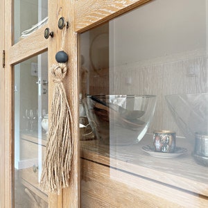 May include: A wooden cabinet with glass doors and shelves. The cabinet has a brown tassel hanging from a knob on the door. The shelves are filled with various items, including a glass bowl, a glass pitcher, and a cup.