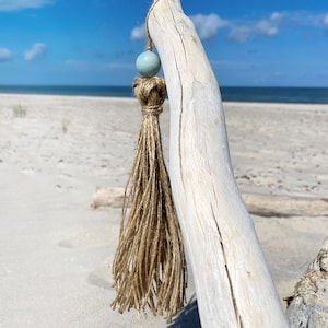 Könnte beinhalten: Eine hellblaue Perle hängt an einem langen, natürlichen Fadenquast. Der Quast ist an einem Stück Treibholz befestigt, das weiß gebleicht ist. Der Hintergrund ist ein Sandstrand mit blauem Himmel und Meer.
